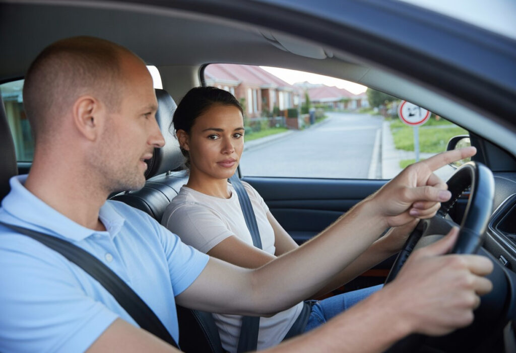 A learner driver and instructor sitting in a car on a suburban street, with the instructor pointing and explaining something to the learner.