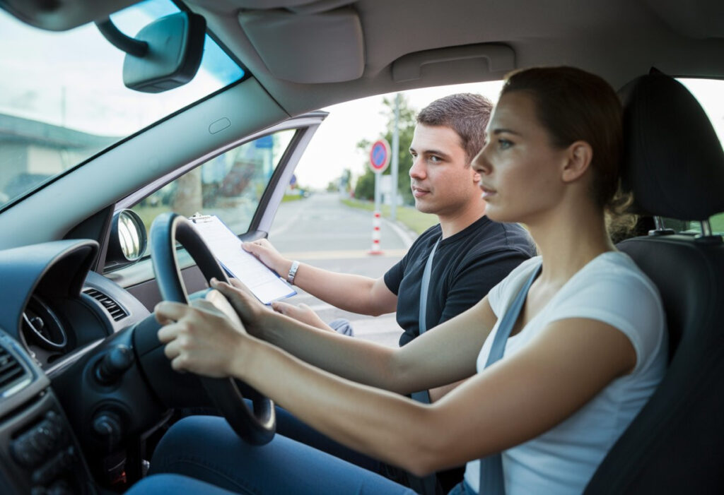 A young adult driver and a driving examiner sitting inside a car during a practical driving test on a suburban street.